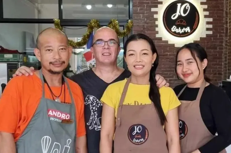 Four smiling employees standing in front of a bakery shop. From left to right: a man in an orange shirt and gray apron, a man with glasses wearing reindeer antlers, a woman in a yellow shirt and brown apron, and another woman in a black shirt and brown apron. Behind them is the bakery's logo on the wall.